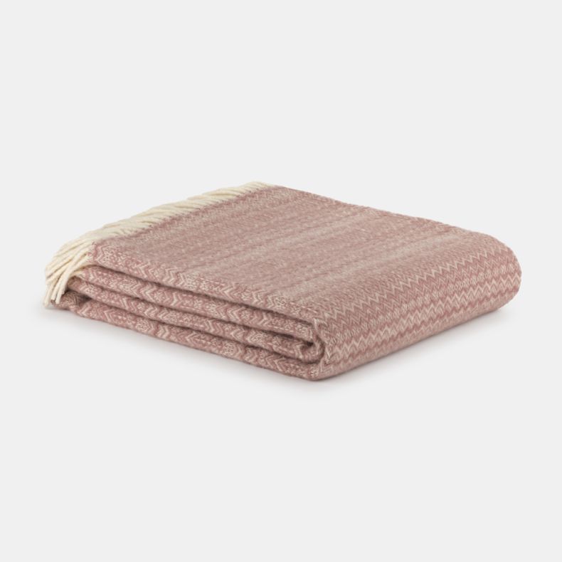 Folded blush pink throw blanket with a soft, cosy woven texture, neatly stacked on a light grey background. Finished with cream fringe tassels and a subtle herringbone-inspired pattern, it offers a warm and inviting look for sofas, beds or armchairs.