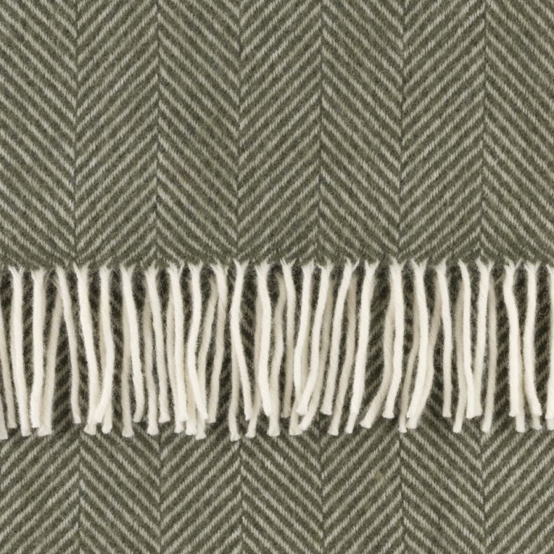 Close-up of a sage green throw blanket featuring a classic herringbone weave and soft cream tassel fringe. The cosy woven texture highlights the timeless chevron-inspired pattern, adding warmth, depth and elegant styling to any living space.