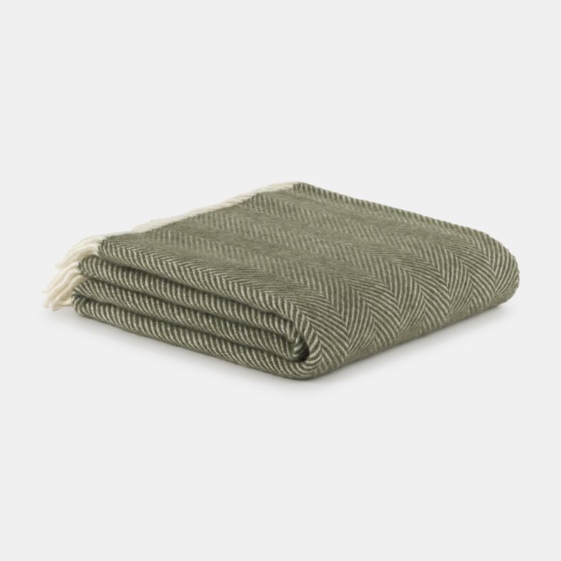 Folded sage green throw blanket with a classic herringbone weave, neatly stacked on a light grey background. Soft and cosy in appearance, the blanket features subtle cream fringe tassels and a timeless textured design, ideal for adding warmth and style to