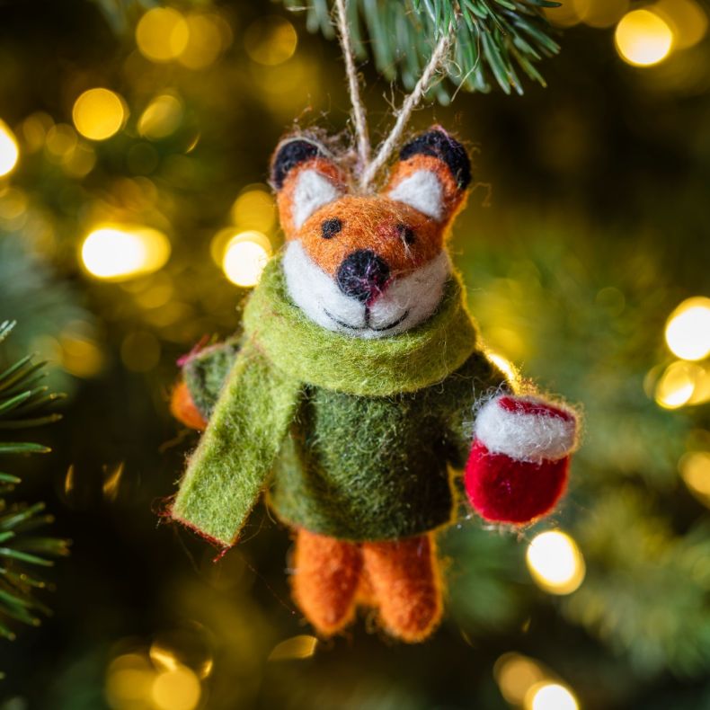 Felt fox Christmas hanging decoration wearing a dark green coat and light green scarf, holding a red mug, featuring an orange and white wool-effect body, needle-felted detailing, and a rustic twine hanging loop, displayed on a Christmas tree among warm wh