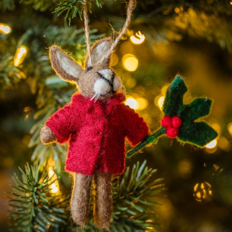 Felt hare Christmas hanging decoration wearing a bright red coat and holding a holly sprig with red berries, featuring a grey-brown wool-effect body, long upright ears, needle-felted detailing, and a rustic twine hanging loop, displayed on a Christmas tre