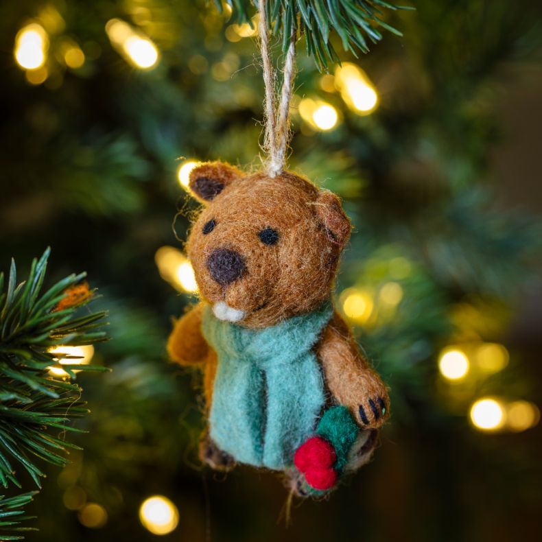 Felt beaver Christmas hanging decoration wearing a pale blue scarf and holding a holly sprig with red berries, featuring a brown wool-effect body, embroidered facial detailing, handcrafted needle-felted finish, and a rustic twine hanging loop, displayed o