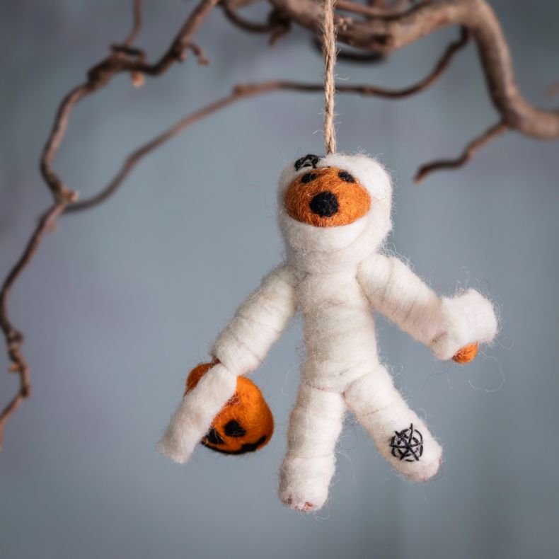 Felt fox mummy hanging decoration crafted from white wool, wrapped in mummy-style bandages and featuring an orange fox face with embroidered black details, holding a small pumpkin lantern and suspended from natural twine against a soft blue background wit