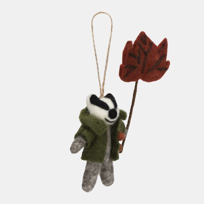 Felt badger hanging decoration shown from the side, featuring a grey felt body, distinctive black-and-white facial markings, and a green coat with orange button details. Holding a red-brown autumn leaf on a stem, this charming seasonal ornament is finishe
