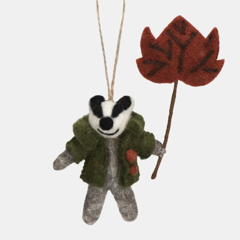 Felt badger hanging decoration wearing a green coat with orange button details and holding a large red-brown autumn leaf on a stem. Crafted with a grey felt body, black-and-white facial markings, and a natural hanging loop, making it a charming seasonal o