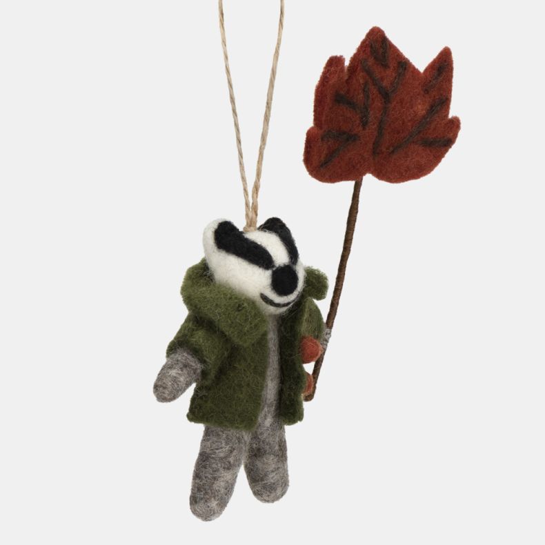 Felt badger hanging decoration shown from the side, featuring a grey felt body, distinctive black-and-white facial markings, and a green coat with orange button details. Holding a red-brown autumn leaf on a stem, this charming seasonal ornament is finishe