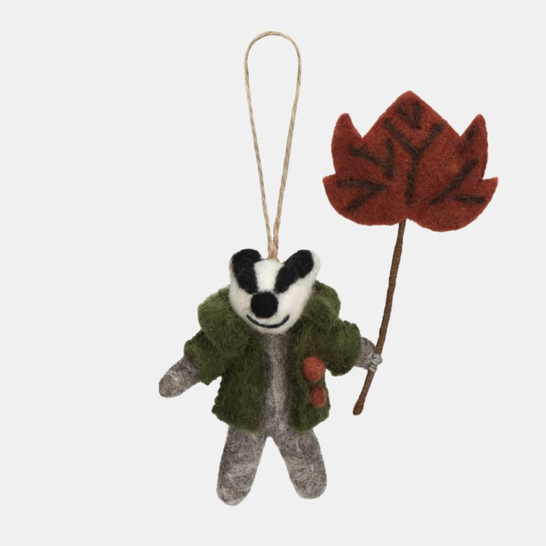 Felt badger hanging decoration wearing a green coat with orange button details and holding a large red-brown autumn leaf on a stem. Crafted with a grey felt body, black-and-white facial markings, and a natural hanging loop, making it a charming seasonal o