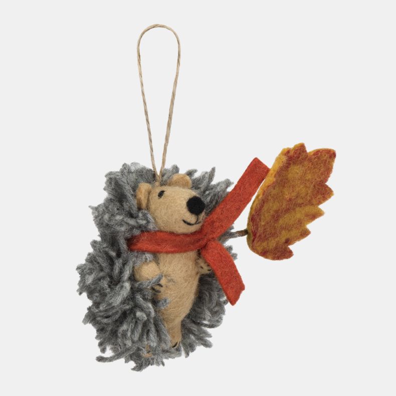 Felt hedgehog hanging decoration shown from the side, featuring a soft grey textured coat, beige felt body, and a bright orange scarf. Holding a large orange and yellow autumn leaf, this charming seasonal ornament is finished with a natural hanging loop a