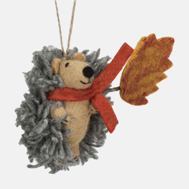 Felt hedgehog hanging decoration shown from the side, featuring a soft grey textured coat, beige felt body, and a bright orange scarf. Holding a large orange and yellow autumn leaf, this charming seasonal ornament is finished with a natural hanging loop a