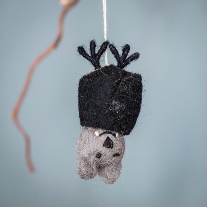 Felt bat hanging decoration crafted from grey and black wool, featuring embroidered facial details, small fangs, raised wings and a hanging string, suspended against a soft blue background.