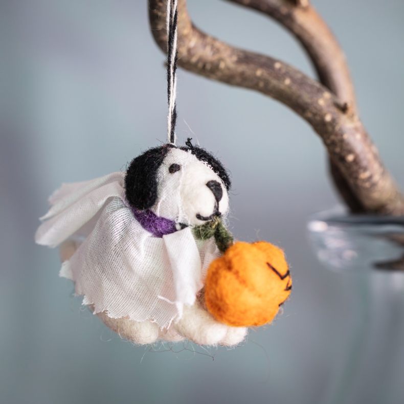 Felt ghost dog hanging decoration crafted from soft wool, featuring a black-and-white dog wrapped in a white ghost costume with a purple collar, holding an orange pumpkin trick-or-treat bucket, suspended from hanging string against a soft blue background 
