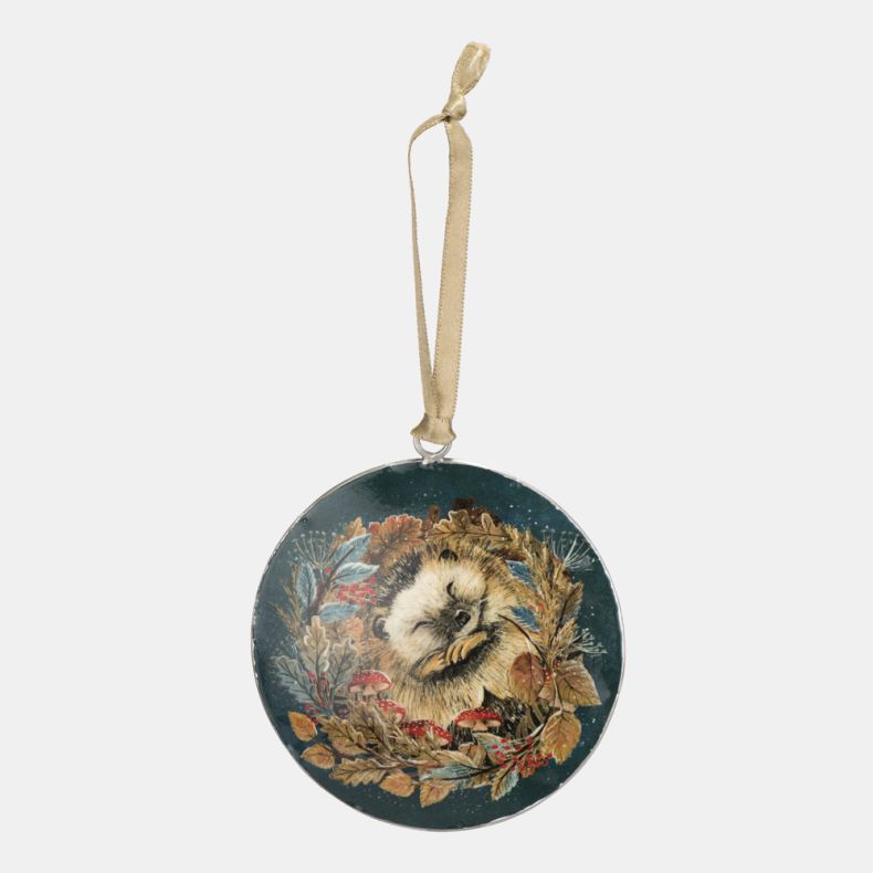 Round hanging Christmas decoration featuring an illustrated sleeping hedgehog surrounded by autumn leaves, berries and woodland mushrooms on a deep teal background, finished with a champagne-coloured ribbon for festive seasonal décor.