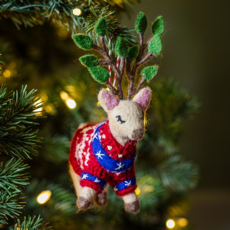 Felt hanging reindeer Christmas decoration displayed on a Christmas tree, featuring green leaf-tipped antlers, embroidered closed-eye detailing, and a red knitted jumper with blue and white festive pattern accents, surrounded by warm white fairy lights an