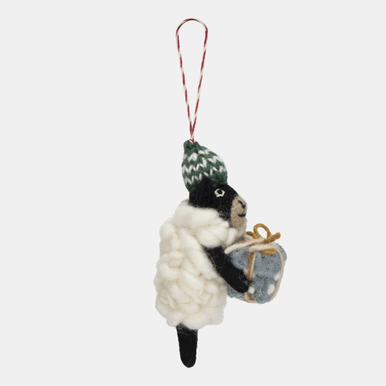 Felt sheep Christmas hanging decoration shown from a side view, featuring a fluffy white wool-effect coat, black face and legs, a green knitted bobble hat with white patterned detailing, and a wrapped blue gift tied with twine, suspended from a red and wh
