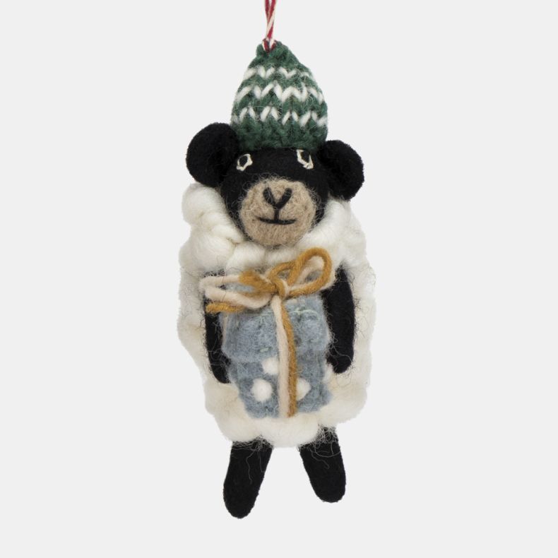 Felt sheep Christmas hanging decoration shown in a close-up view, featuring a fluffy white wool-effect coat, black face and legs, a green knitted bobble hat with white patterned detailing, and a pale blue gift wrapped with twine, suspended from a red and 