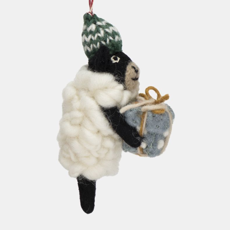 Felt sheep Christmas hanging decoration shown from a close-up side view, featuring a fluffy white wool-effect coat, black face and legs, a green knitted bobble hat with white patterned detailing, and a pale blue gift wrapped with twine, suspended from a r