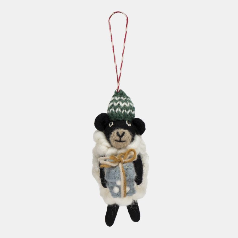 Felt sheep Christmas hanging decoration featuring a fluffy white wool-effect coat, black face and legs, a green knitted bobble hat with white patterned detailing, and a wrapped blue gift tied with twine, suspended from a red and white hanging loop.