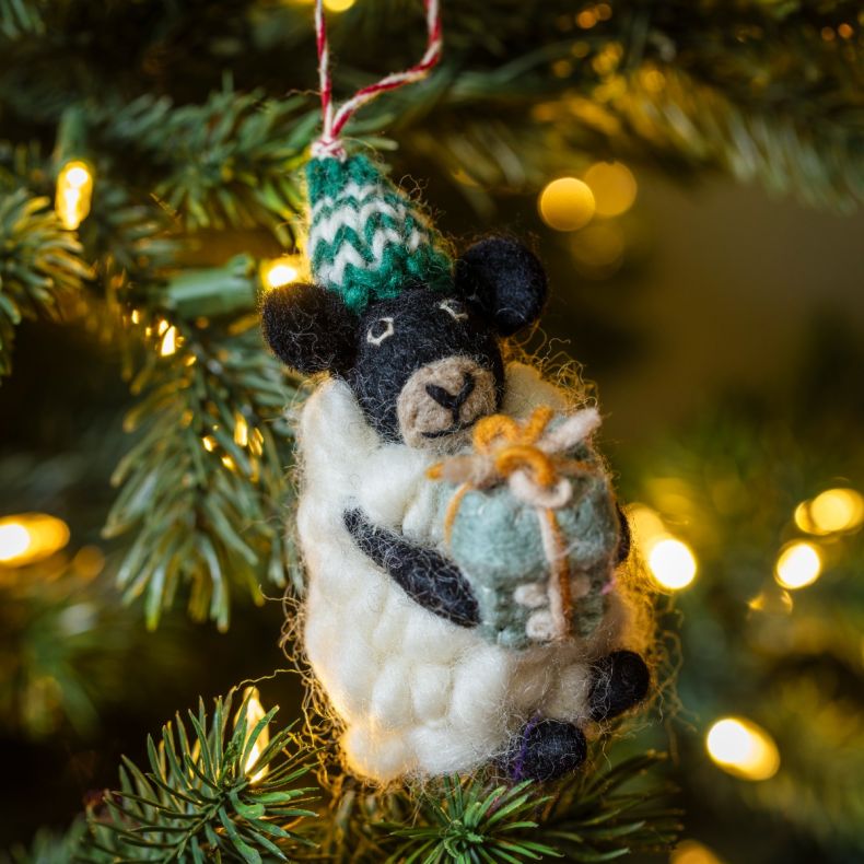 Felt sheep Christmas hanging decoration displayed on a Christmas tree, featuring a fluffy white wool-effect coat, black face and legs, a green knitted bobble hat with white patterned detailing, and a pale blue gift wrapped with twine, surrounded by warm w