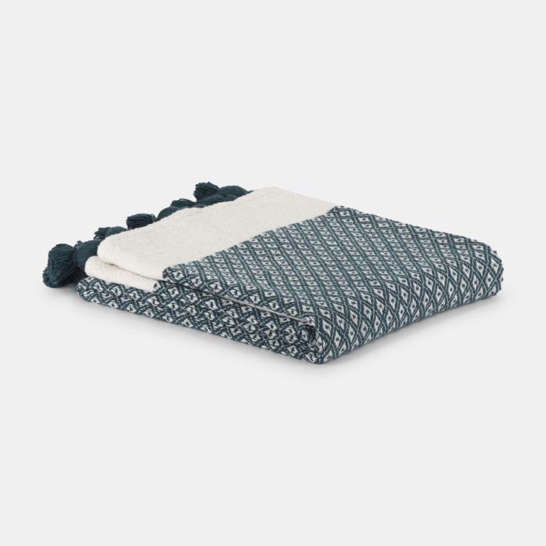 Folded woven throw blanket in teal and cream, featuring a geometric diamond pattern and decorative teal tassels along the edge. Soft textured fabric displayed neatly folded against a light grey background.