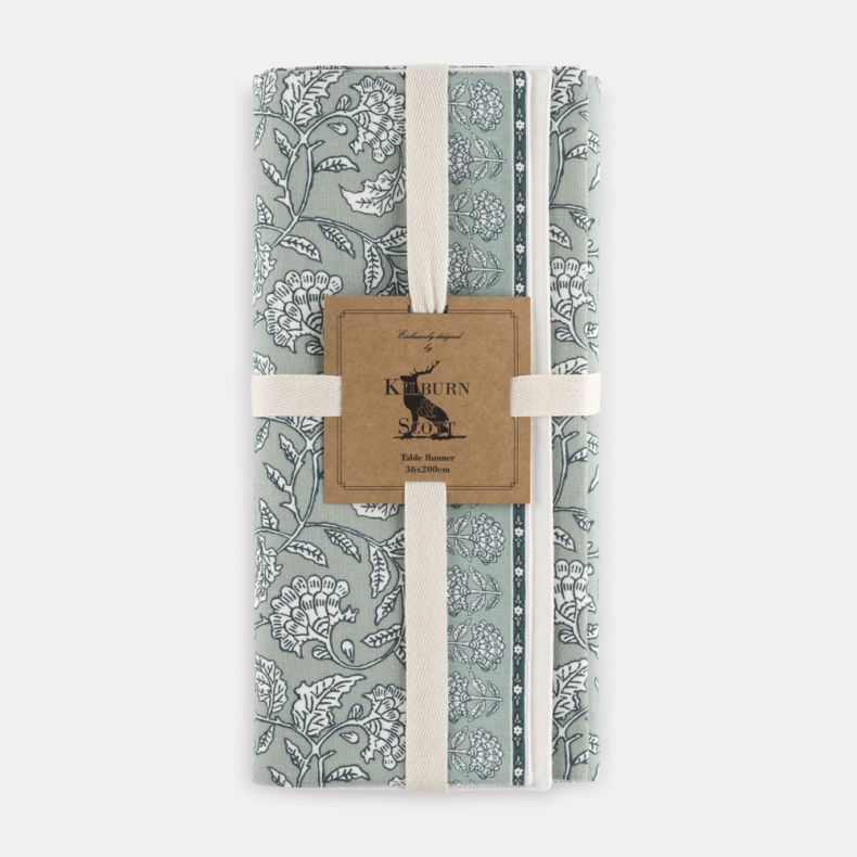 Kilburn & Scott Table Runner in soft sage green, featuring an elegant heritage-inspired floral and bird print with decorative border detailing. Folded and secured with presentation straps, this fabric table runner combines classic country charm with a mut