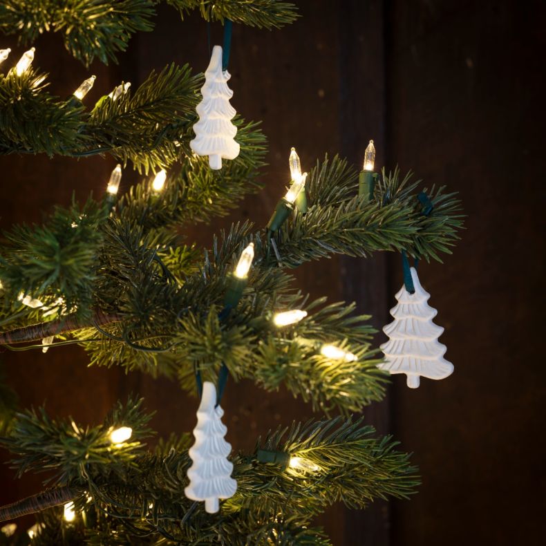 White ceramic Christmas tree decorations hanging on a lit Christmas tree, featuring embossed branch detailing, teal ribbon loops, and a festive Frosted Pines fragrance design displayed among warm white fairy lights and evergreen branches.