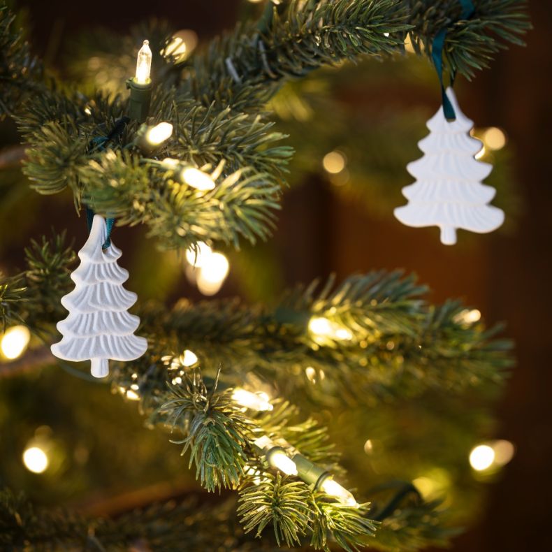 White ceramic Christmas tree decorations hanging on a lit Christmas tree, featuring embossed branch detailing, teal ribbon loops, and a festive Frosted Pines fragrance design among warm white fairy lights and evergreen branches.