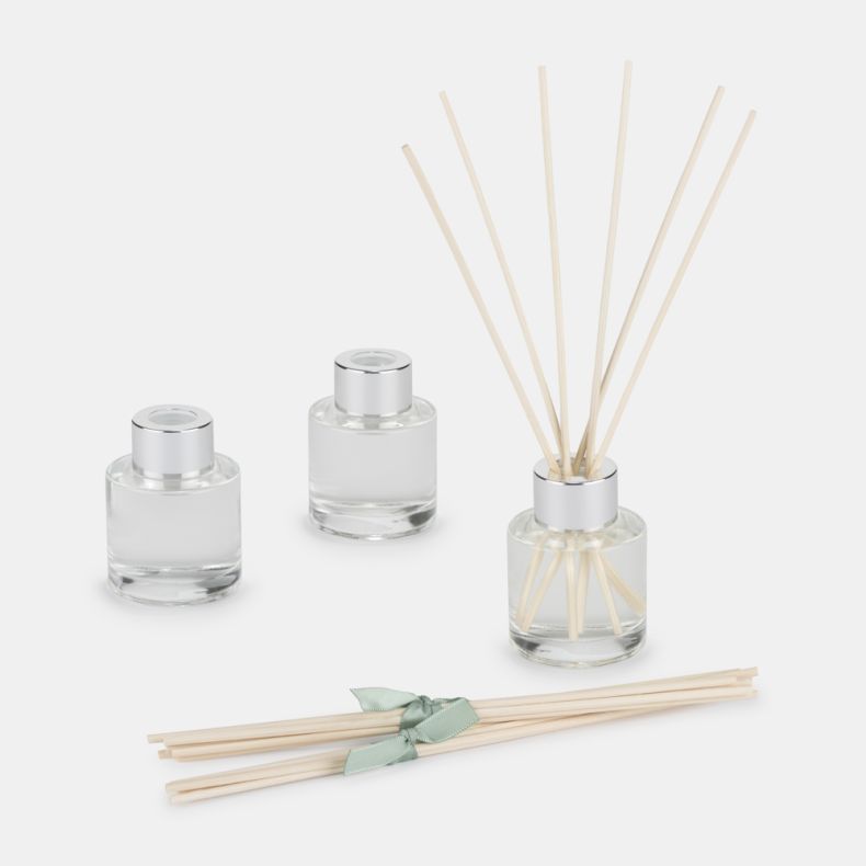 Luxury reed diffuser set with three clear glass fragrance bottles, silver caps, natural reed sticks, and a bundle of spare reeds tied with a soft green ribbon. Displayed on a light background, ideal for home fragrance gifting.