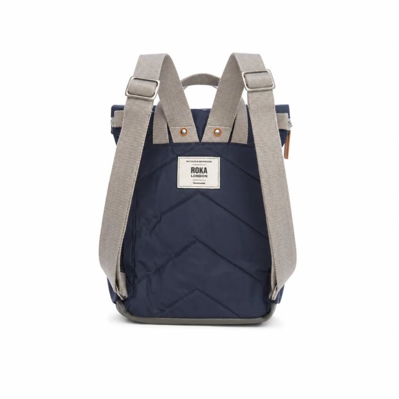 Roka London backpack shown from the back in navy blue, featuring adjustable light grey shoulder straps, quilted back panel and top carry handle. Lightweight design with a rear Roka London logo patch and durable water-resistant finish.