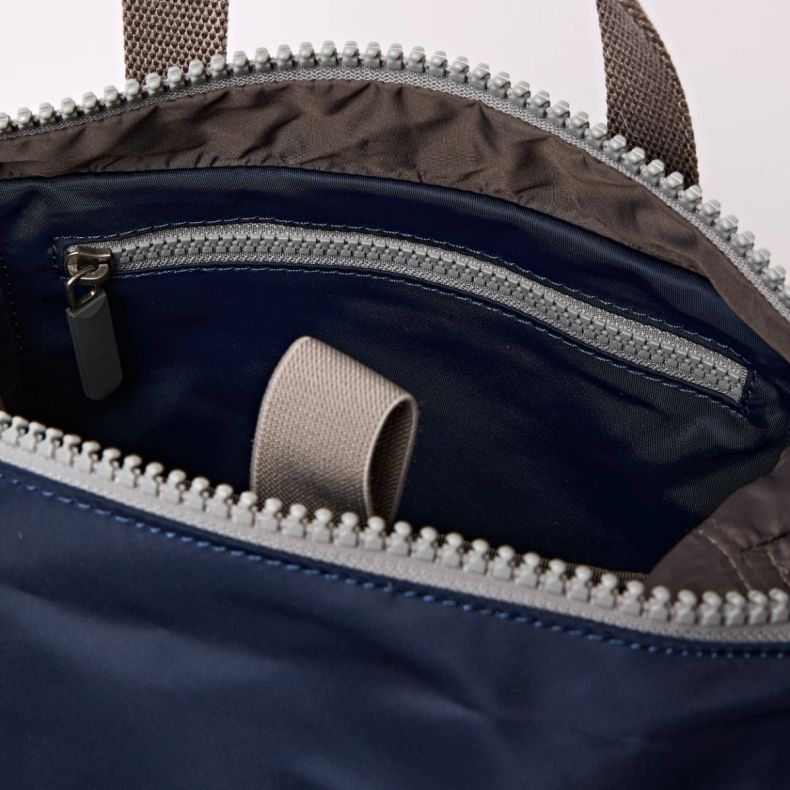 Close-up view of the Roka London backpack interior, showing a navy lining, internal zip pocket, wide opening with grey zip fastening, and a fabric carrying loop inside. The organised compartment design offers practical storage for everyday essentials.