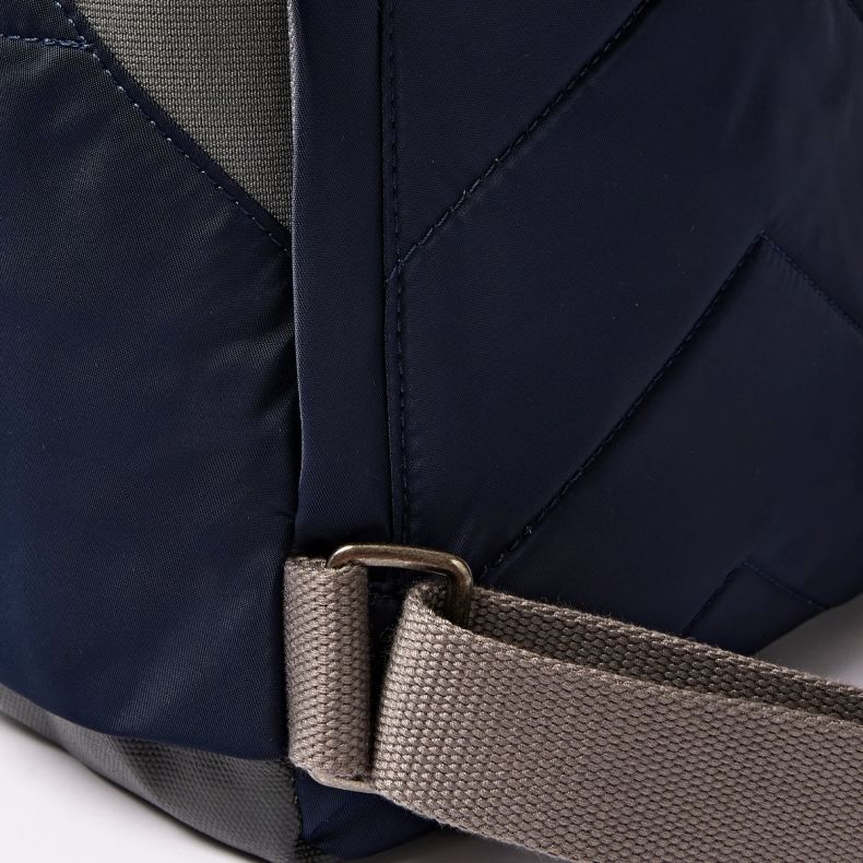 Close-up detail of the Roka London backpack back panel, showing navy blue quilted water-resistant fabric, a grey adjustable shoulder strap and metal hardware. The image highlights the bag’s durable construction and practical design features.