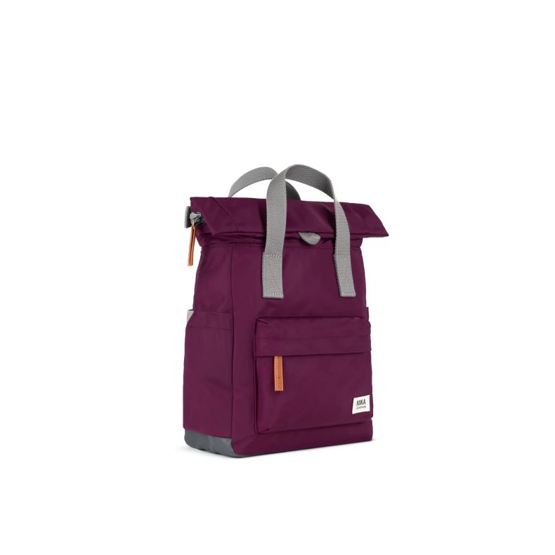 Angled view of a burgundy red Roka London backpack with a roll-top closure, grey webbing carry handles, front zip pocket, side pockets, and tan zip pulls. The lightweight backpack features a minimalist design and Roka London logo label on the front.
