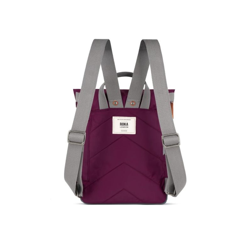 rear view of a burgundy red Roka London backpack featuring adjustable grey shoulder straps, padded back panel with stitched chevron detailing, top carry handles, and a woven Roka London logo label. The roll-top backpack is shown against a plain light back