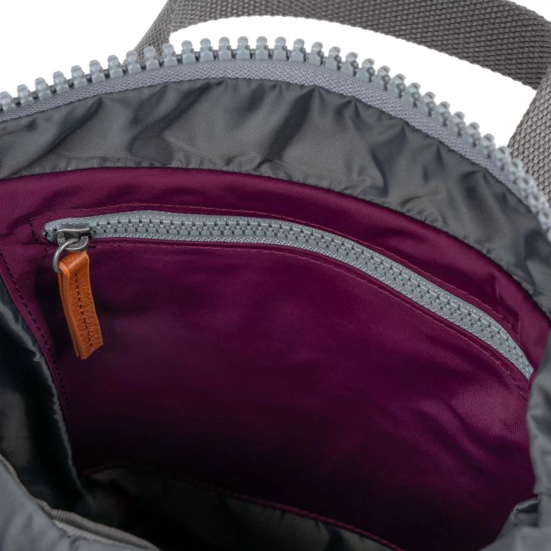 Close-up view of the interior of a burgundy red Roka London backpack, showing a grey lined compartment with an internal zip pocket. The pocket features a grey zipper and tan pull tab, highlighting the organised storage space and durable lining.