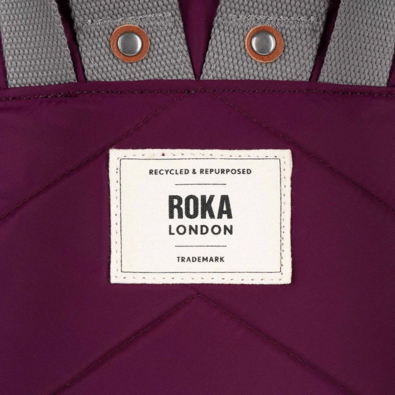 Close-up of the back panel of a burgundy red Roka London backpack, featuring a stitched chevron pattern, grey webbing straps with metal rivets, and a woven Roka London logo patch made from recycled and repurposed materials.