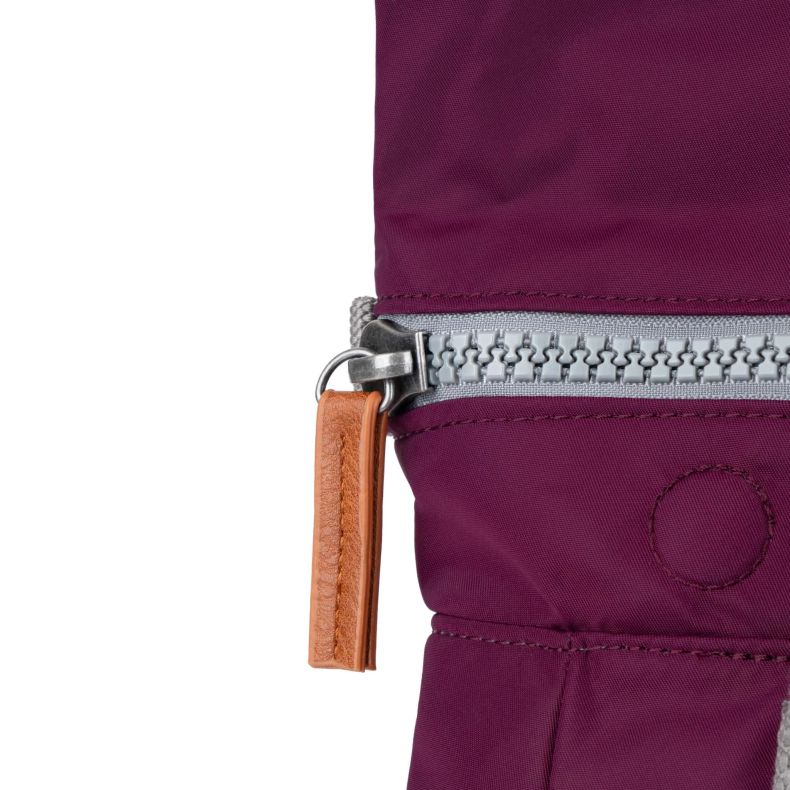Close-up of a burgundy red Roka London backpack showing a grey zipper with oversized teeth and a tan leather-look pull tab. The detailed view highlights the durable water-resistant fabric, stitching, and hardware used in the backpack’s construction.