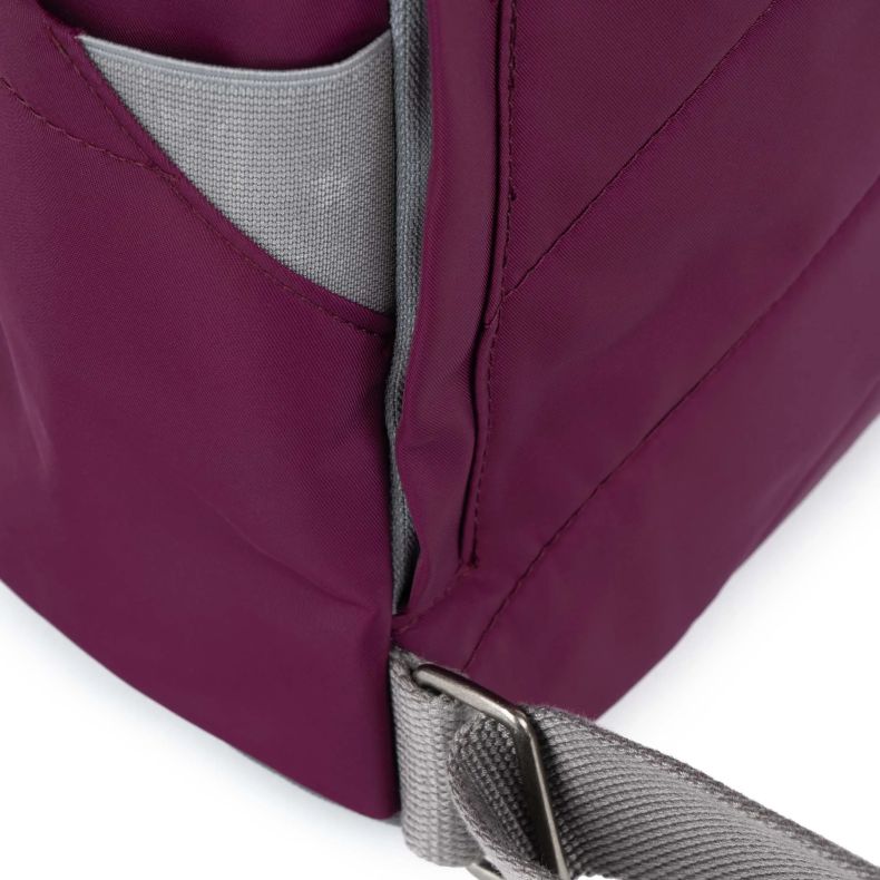 Close-up of a burgundy red Roka London backpack showing a grey side pocket, adjustable grey shoulder strap, and reinforced stitching. The detailed view highlights the durable water-resistant fabric, practical storage design, and sturdy metal strap hardwar
