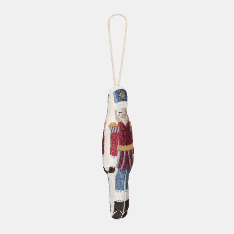 Embroidered nutcracker soldier Christmas hanging decoration shown from an angled side view, featuring a red military-style jacket, blue trousers, white hat with gold star detail, stitched facial features, decorative embroidered detailing, and a cream hang
