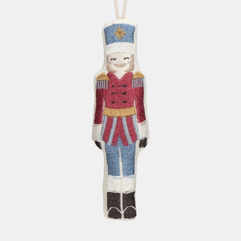 Embroidered nutcracker soldier Christmas hanging decoration shown in a close-up front view, featuring a red military-style jacket with gold detailing, blue trousers, white hat with gold star motif, stitched facial features, decorative embroidery, and a cr