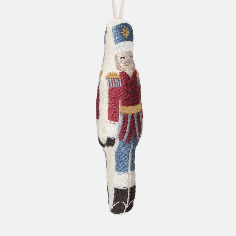 Embroidered nutcracker soldier Christmas hanging decoration shown in a close-up angled side view, featuring a red military-style jacket with gold detailing, blue trousers, white hat with gold star motif, stitched facial features, decorative embroidery, an