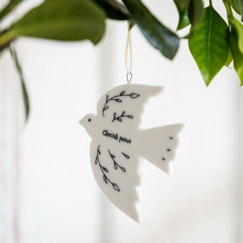 White ceramic dove hanging decoration displayed among green foliage, featuring delicate black botanical line illustrations, the words “Cherish peace”, a smooth glazed finish, and a cream hanging ribbon, shown styled as a festive seasonal ornament.