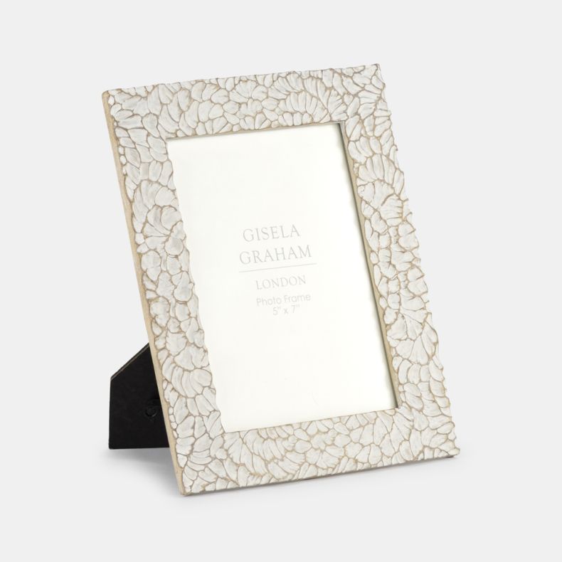 Angled view of a decorative photo frame featuring an intricate shell-inspired mosaic pattern in soft cream tones with subtle distressed detailing. Designed for a 5" x 7" photograph, the textured frame includes a fold-out stand for tabletop display, adding