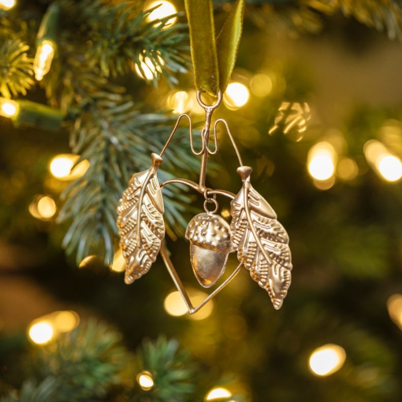Gold-tone hanging acorn decoration displayed on a Christmas tree, featuring detailed oak leaf motifs, a textured acorn charm, geometric wire frame detailing, and an olive green velvet hanging ribbon, shown among evergreen branches and warm white fairy lig