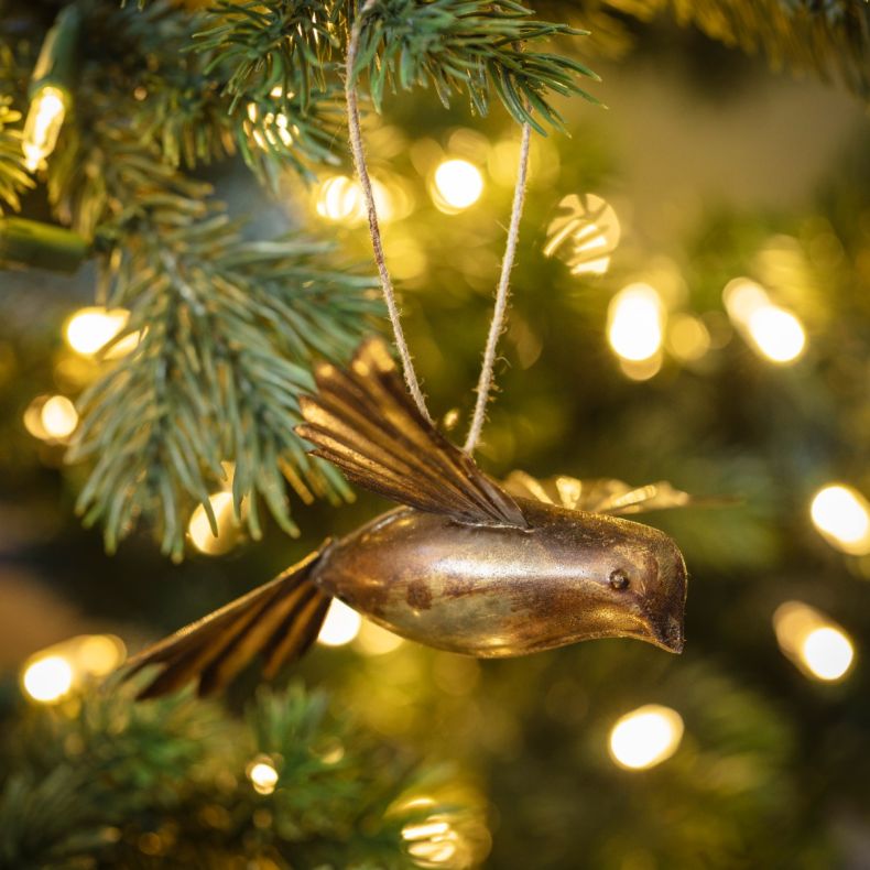 Gold-tone metal bird hanging decoration displayed on a Christmas tree, featuring a sculpted bird design with embossed feather detailing, an antique brass-effect finish, and a rustic twine hanging loop, shown among evergreen branches and warm white fairy l
