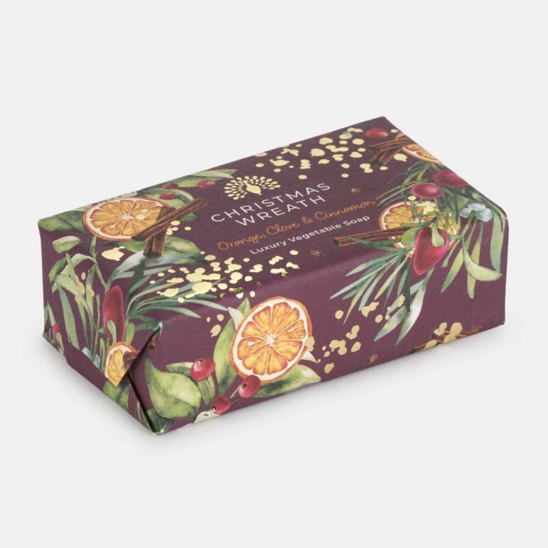 Christmas Wreath luxury vegetable soap in festive gift packaging, shown from an angled view and wrapped in a deep burgundy paper featuring illustrated oranges, cinnamon sticks, berries, foliage and gold accents, scented with orange, clove and cinnamon, is