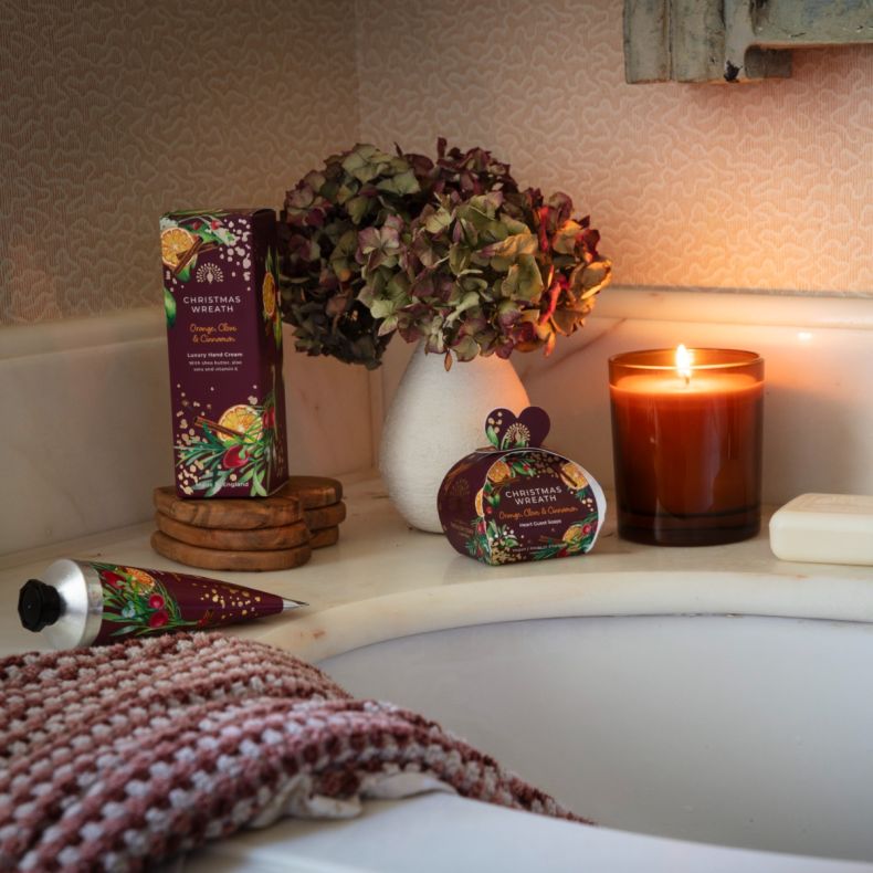 Christmas Wreath hand care and soap gift collection displayed beside a bath, featuring festive orange, clove and cinnamon scented packaging, a luxury hand cream tube, boxed hand cream, heart-shaped guest soaps, a lit candle and a vase of dried hydrangeas 