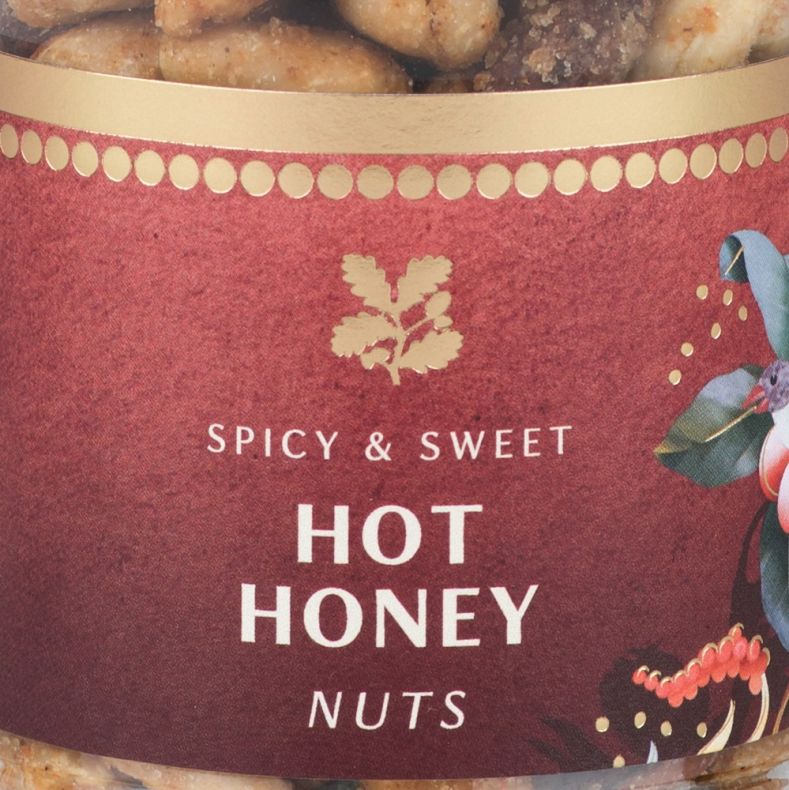 Close-up of a jar of spicy and sweet hot honey nuts, showing a burgundy label with white product lettering, gold decorative accents and botanical artwork, with honey-coated mixed nuts visible through the clear glass jar above and below the label.