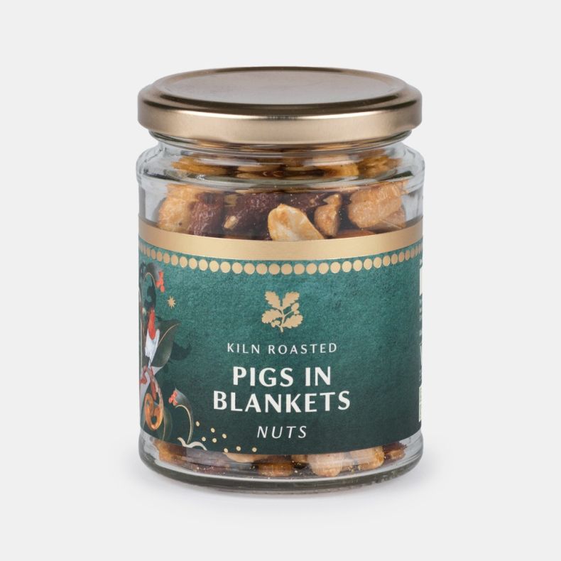 Jar of kiln roasted pigs in blankets nuts, featuring a clear glass jar with a gold lid and a dark green label decorated with festive botanical artwork, showing roasted mixed nuts inside and isolated on a white background.