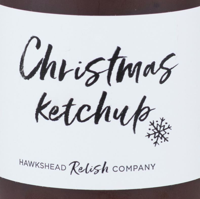 Close-up of a bottle of Christmas ketchup from Hawkshead Relish Company, showing a white label with handwritten-style black lettering, a snowflake illustration and brand logo, wrapped around a glass bottle filled with rich red ketchup