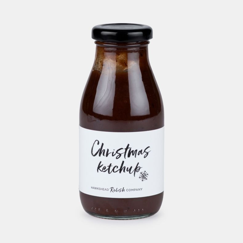 Bottle of Christmas ketchup from Hawkshead Relish Company, featuring a glass bottle with a black lid and a white label with handwritten-style text, filled with rich red ketchup and isolated on a white background.