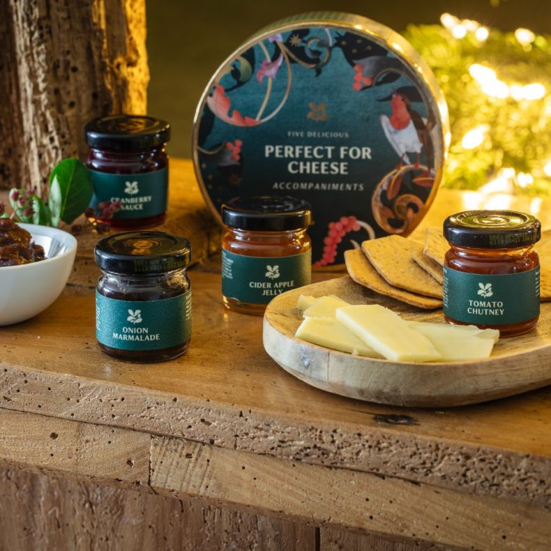 Perfect for Cheese accompaniments gift set featuring miniature jars of onion marmalade, cider apple jelly, tomato chutney and cranberry sauce, displayed alongside sliced cheese and crackers on a rustic wooden table with festive seasonal styling.